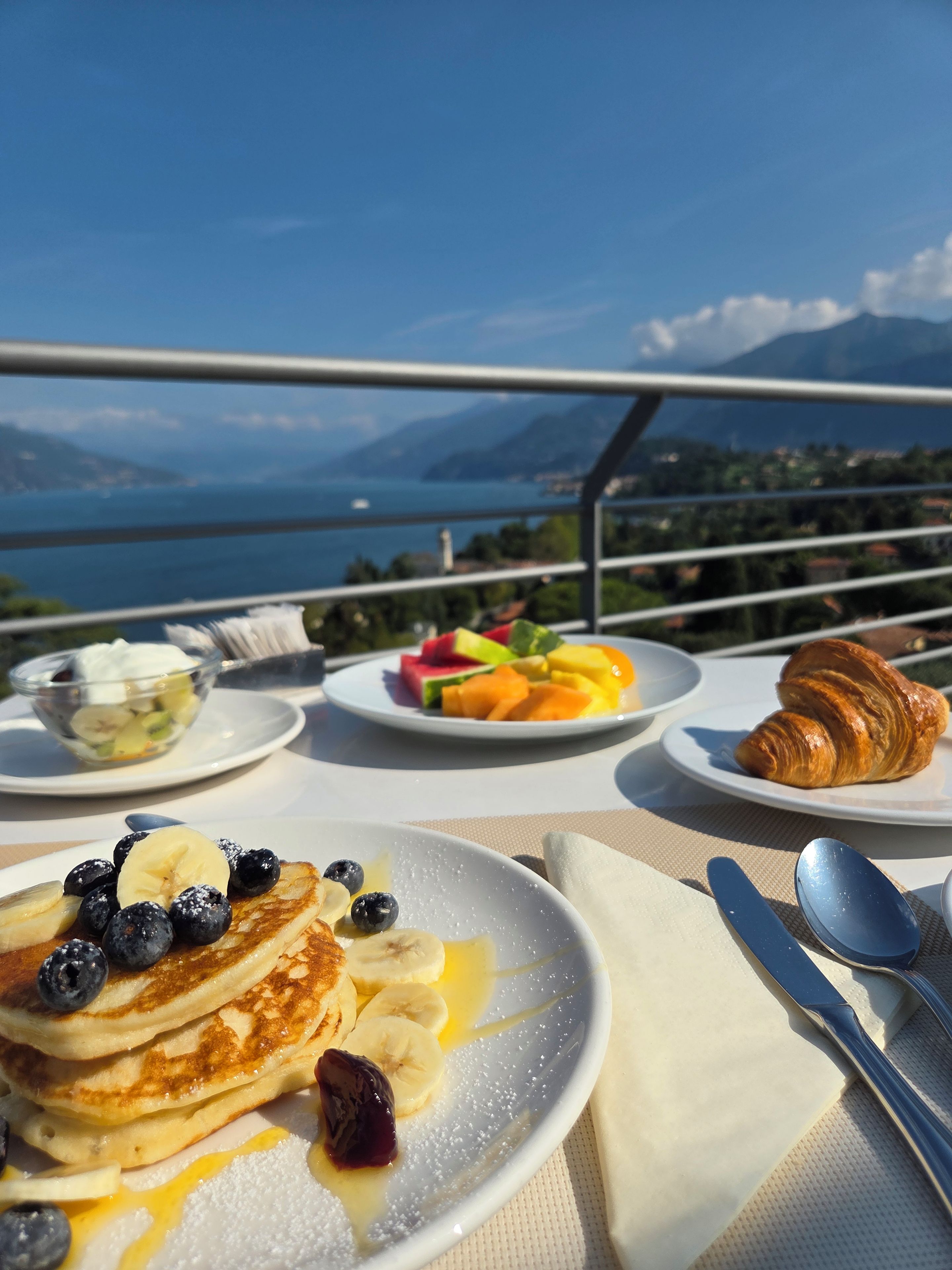 A table with breakfast items including pancakes, fruit, and a croissant, with a scenic view in the background.