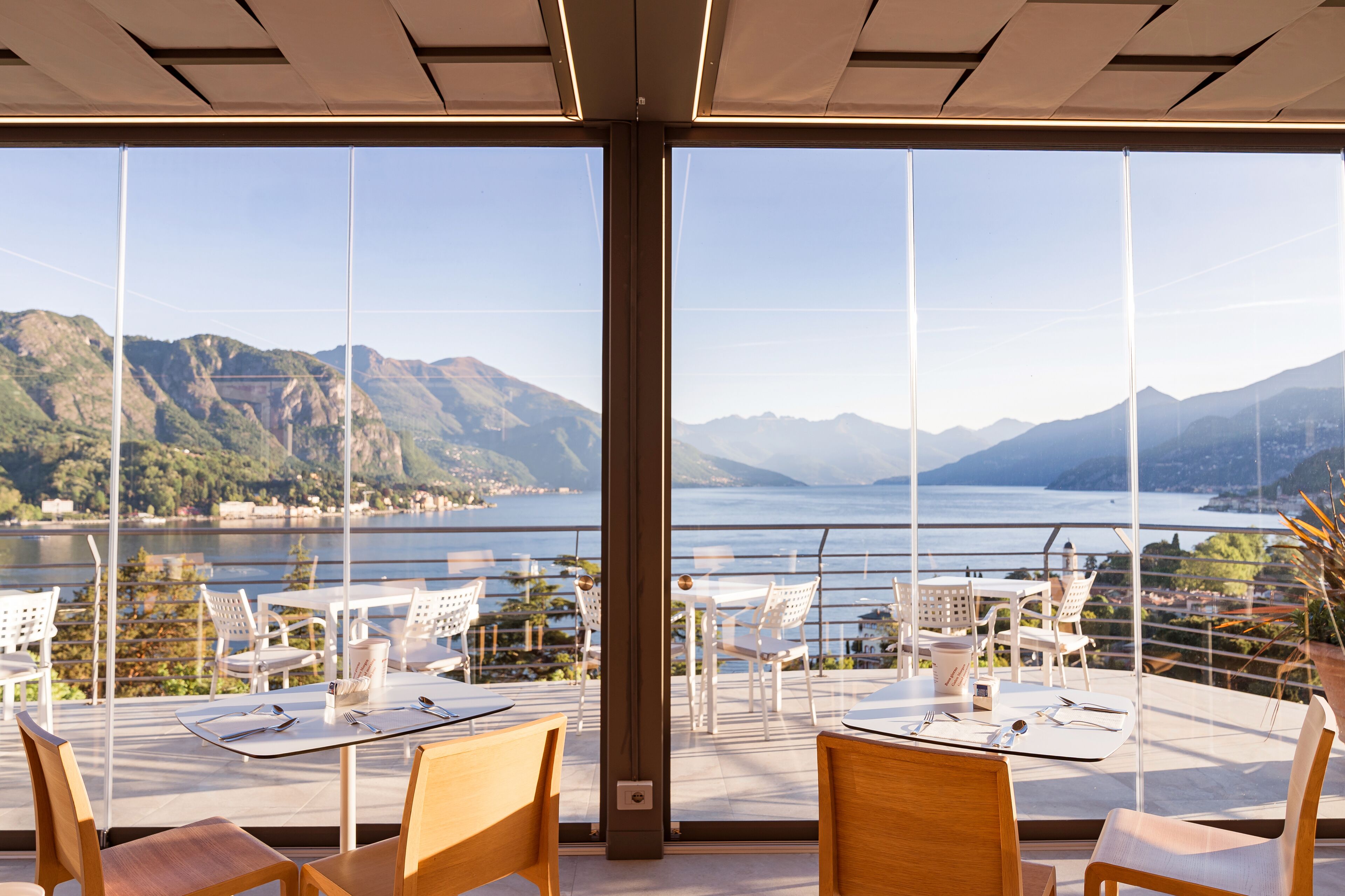 A terrace with tables and chairs, a view of mountains and a lake, and a clear sky.