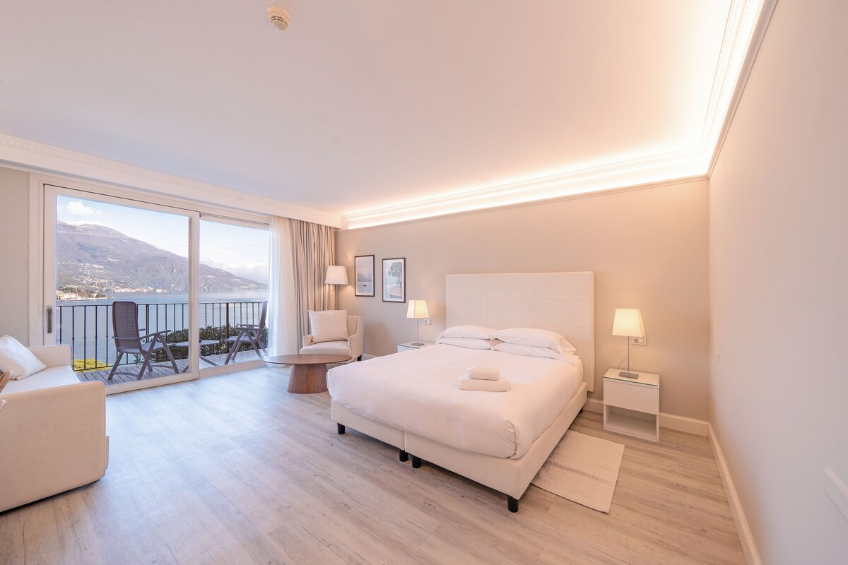 A hotel room with a large bed, a balcony with chairs, a small table, and a view of the mountains.