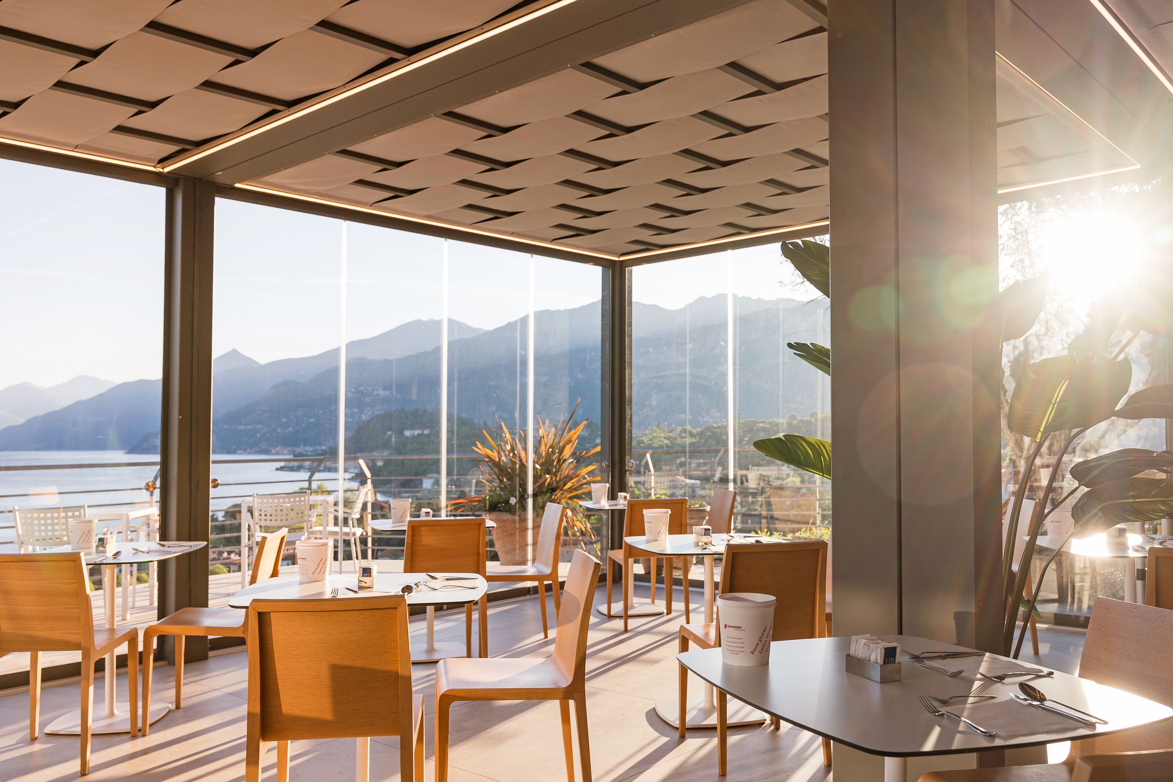 Un espace de restauration moderne, avec des tables et des chaises, de grandes fenêtres offrant une vue sur les montagnes et l’eau, et un plafond orné d’un motif géométrique.