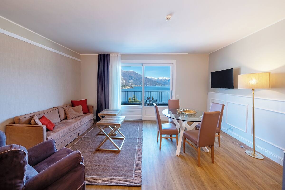 A modern living room with a sofa, a dining table with chairs, a TV mounted on the wall, and a view of the sea through a large window.