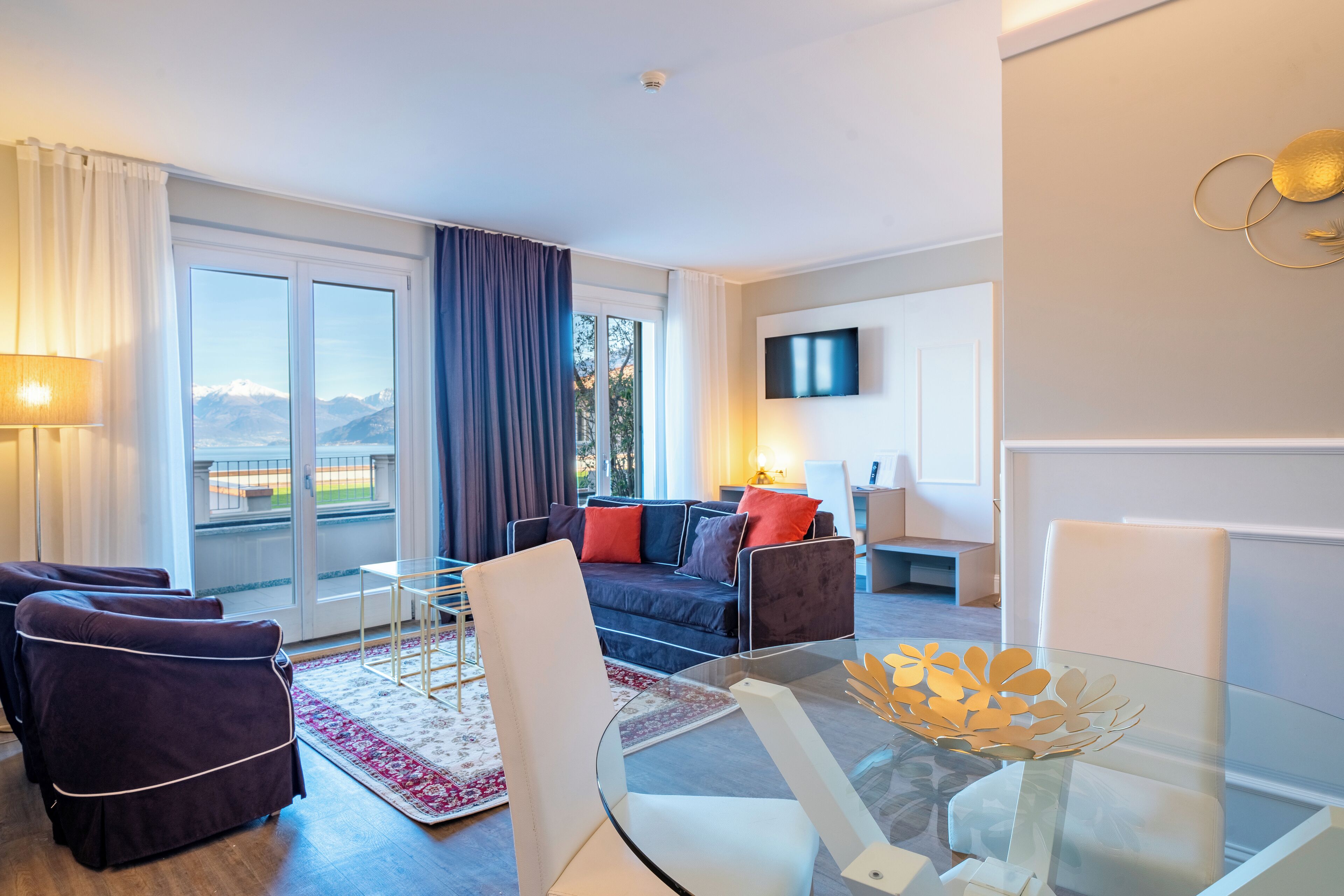 A modern hotel room with a glass table, comfortable seating, a large window with a view, and a flat-screen TV.