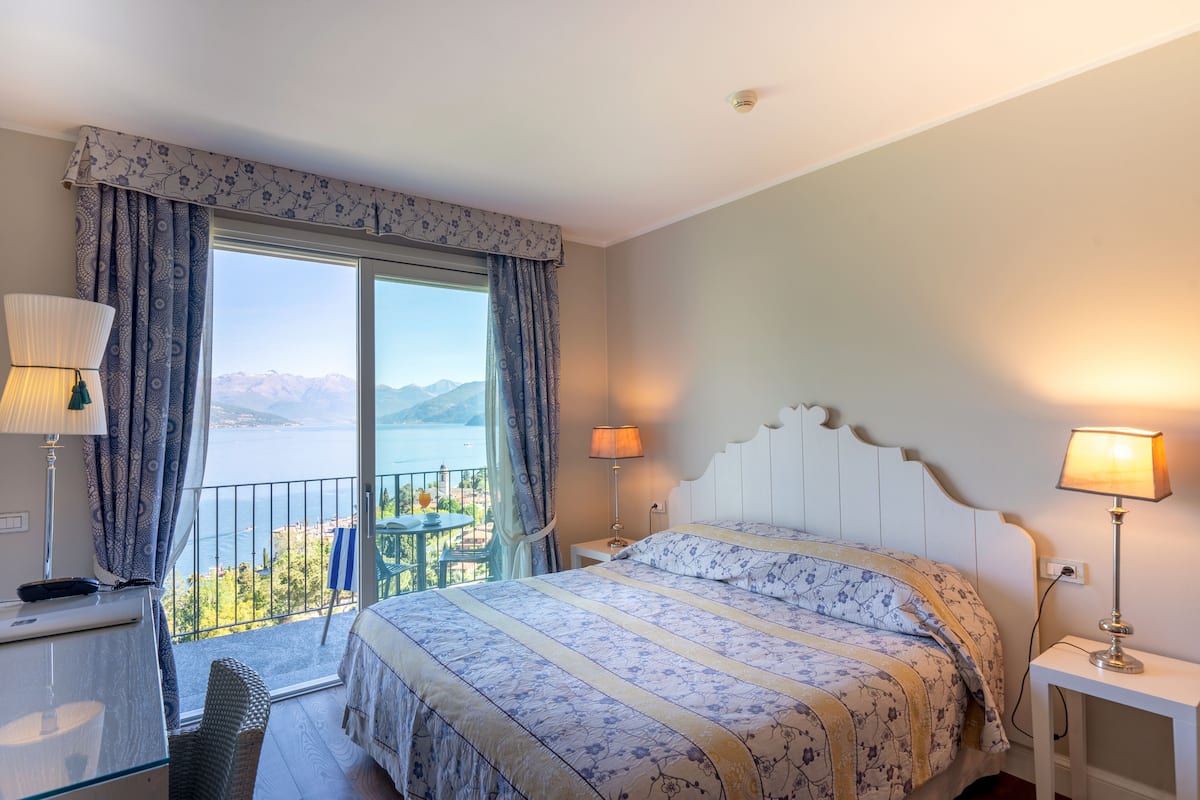 A hotel room with a bed, a desk, a chair, a balcony with a table, and a view of the sea and mountains.