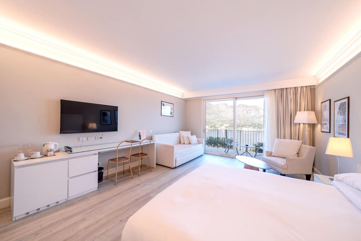 A hotel room with a large bed, a desk with a chair, a sofa, a TV, and a balcony with a view.