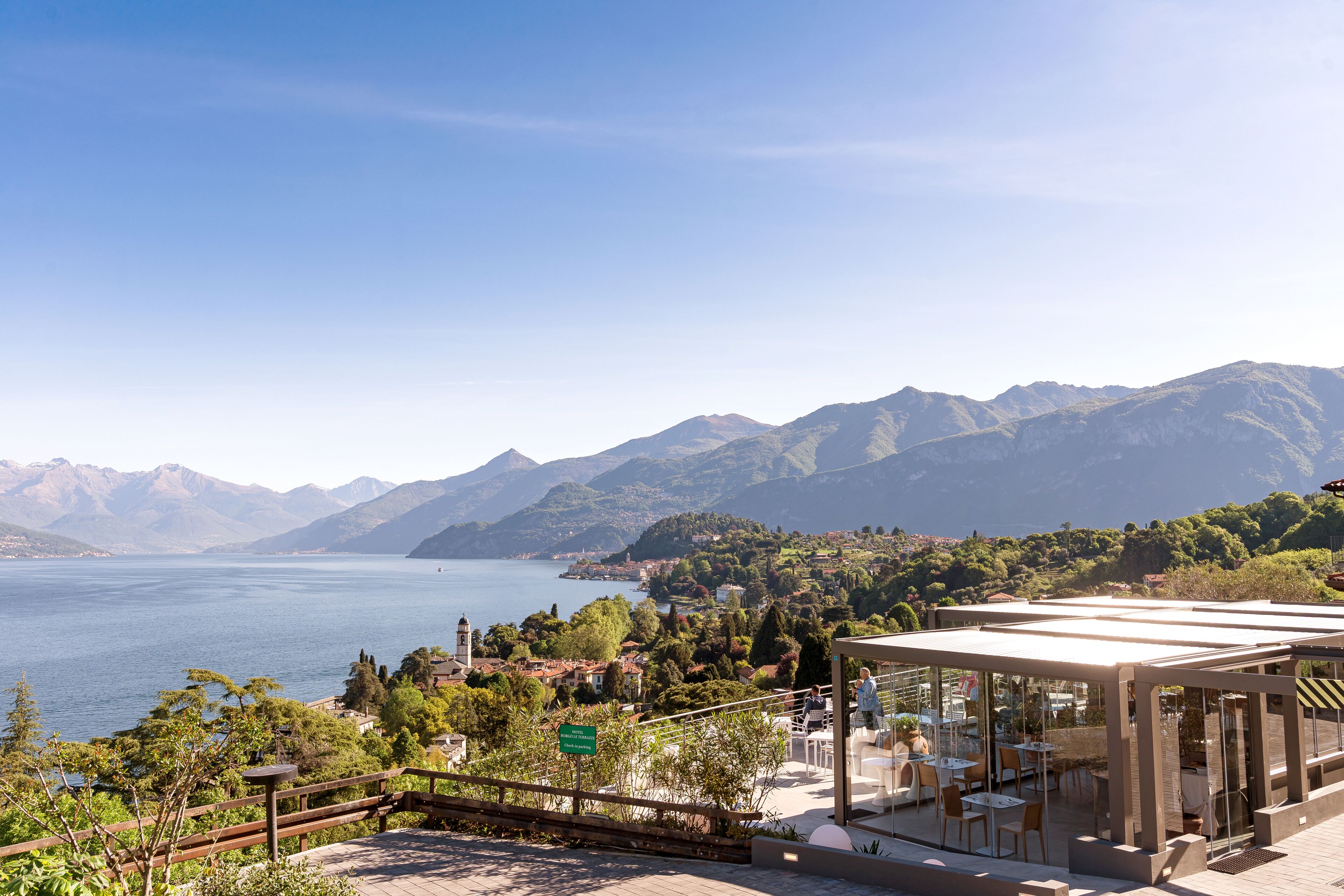 A lakeside terrace with outdoor seating, a mountain backdrop, and a clear sky.