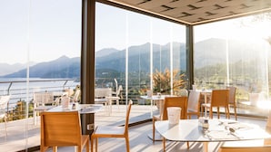 A modern dining area with tables and chairs, large windows with a mountain view, and a glass railing overlooking a body of water. - Borgo Le Terrazze (Bellagio)
