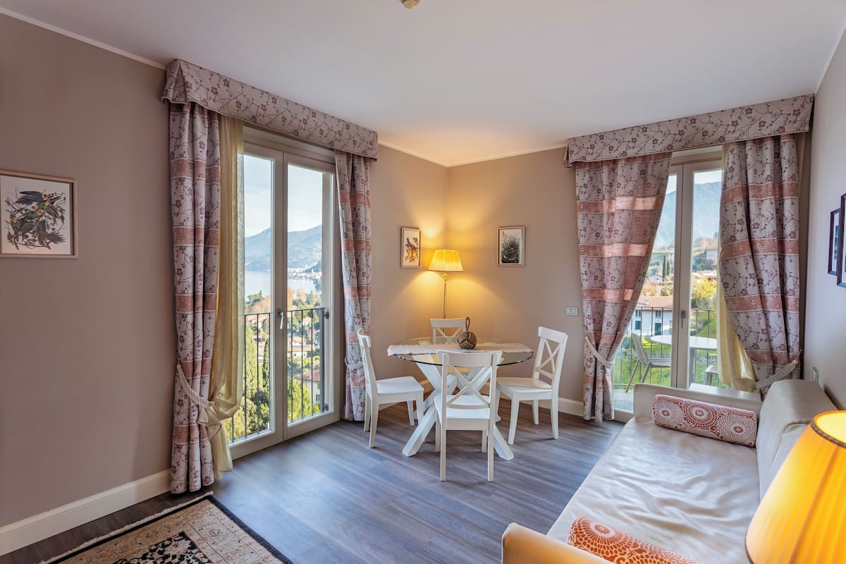 A room with a bed, a table with chairs, a lamp, and a view of the outdoors.