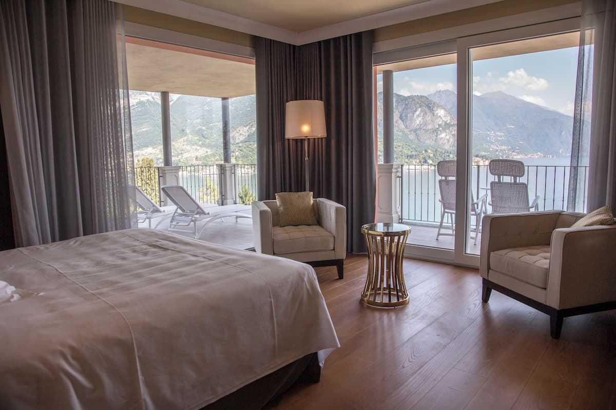 A hotel room with a large bed, two armchairs, a small table, and a balcony with a view of mountains.