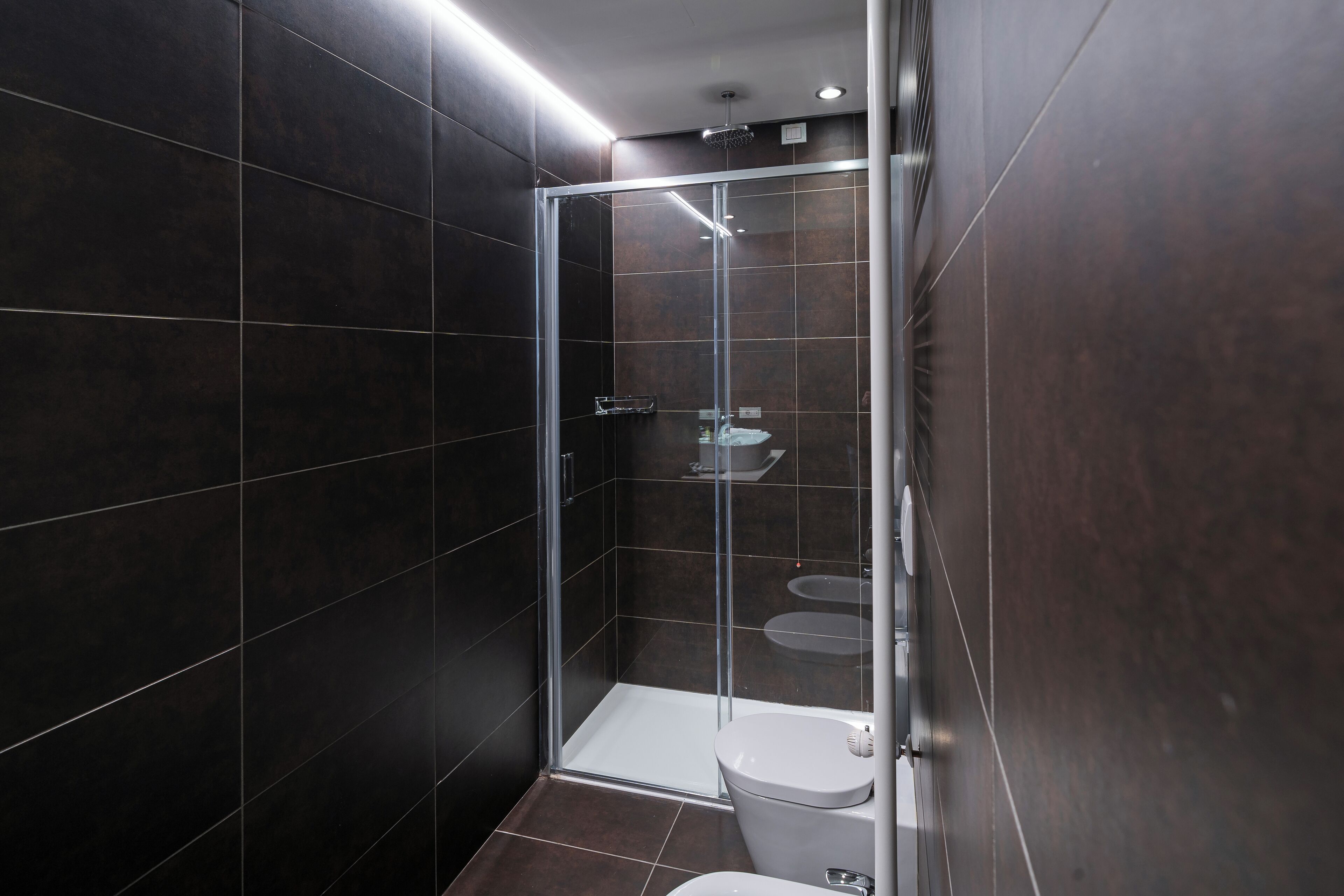 A modern bathroom with dark tiles, a glass-enclosed shower, a toilet, and a sink.