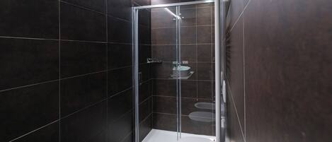 Un baño moderno con azulejos oscuros, ducha con cerramiento de vidrio, inodoro y lavamanos.