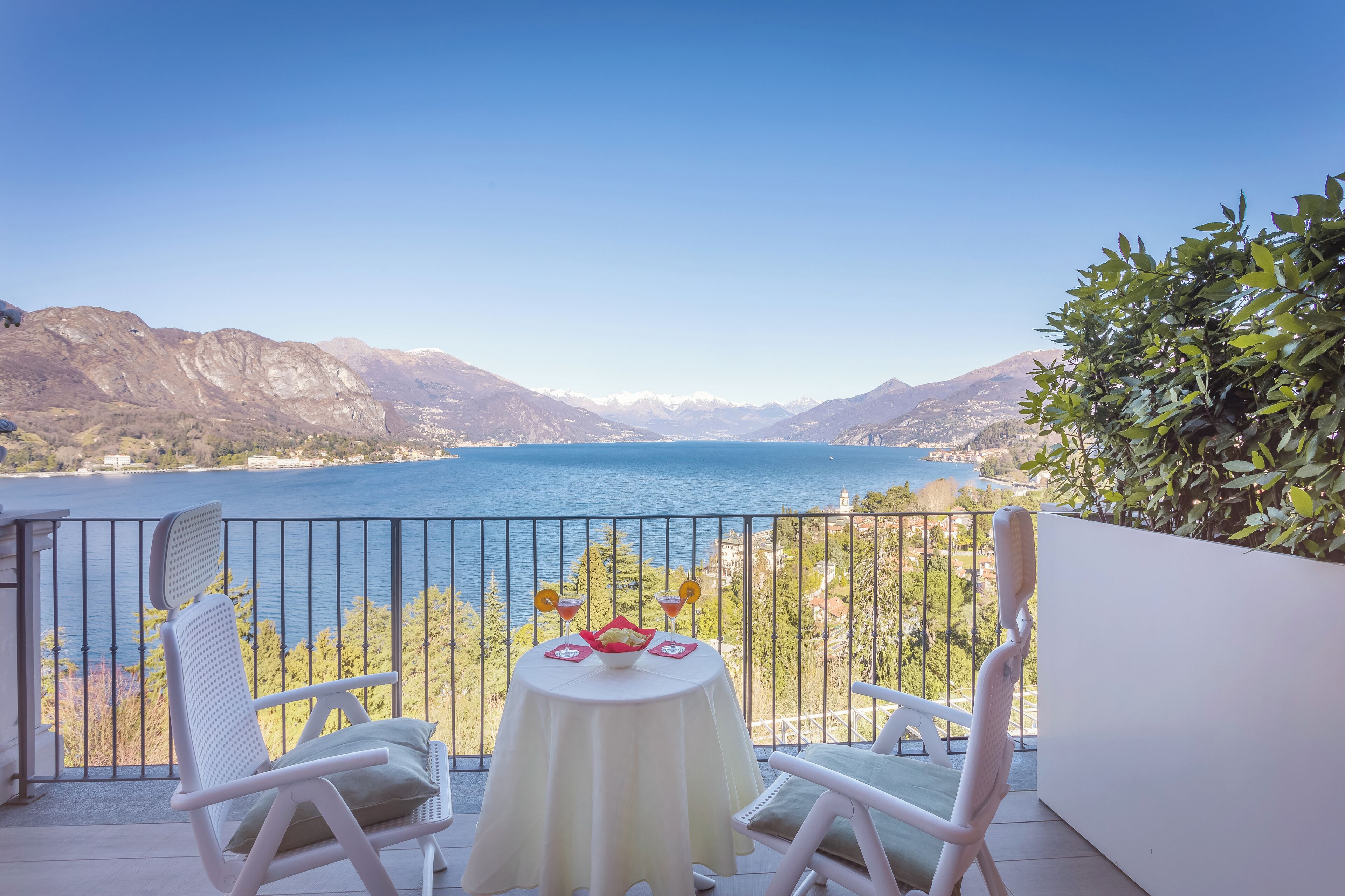 Ein Balkon mit zwei weißen Stühlen, einem Tisch mit weißem Tischtuch und Blick auf einen See und Berge.