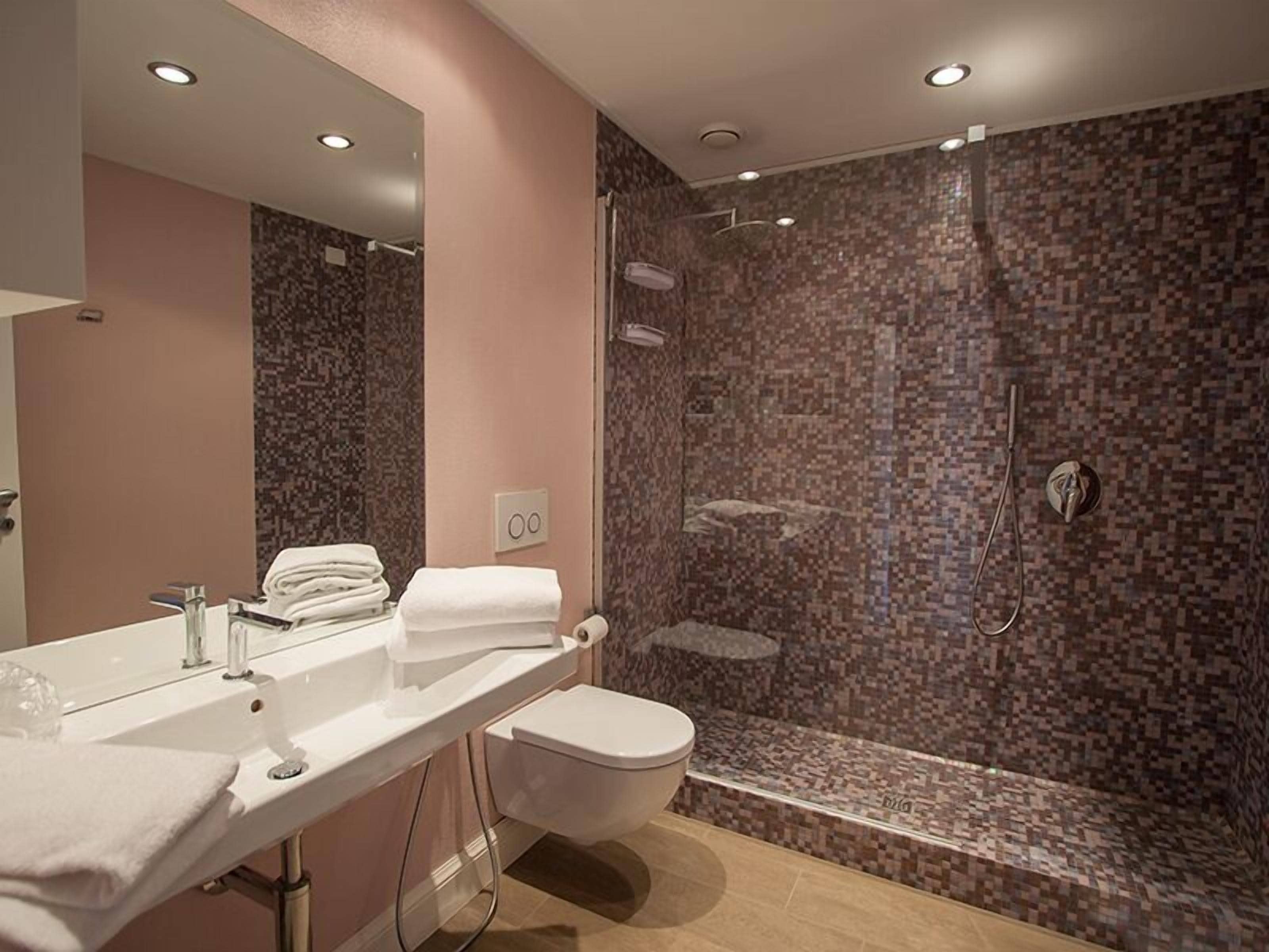 A modern bathroom with a large mirror, a sink, a toilet, and a shower with a mosaic tile wall.