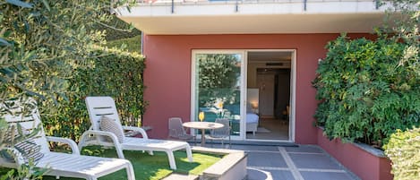 Un balcón con barandillas blancas, un pequeño espacio exterior con sillas y una mesa blancos, y una puerta de cristal que da a un espacio interior.
