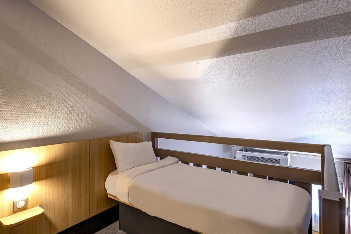 Ein Einzelbett in einem Raum mit schrägverlaufender Decke, einer Wandleuchte und einer Klimaanlage.