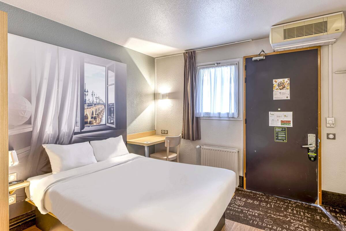 Ein Hotelzimmer mit Bett, Schreibtisch, Stuhl, einem Fenster mit Vorhängen, einer Klimaanlage und einer Tür mit Schloss und Hinweisschild.