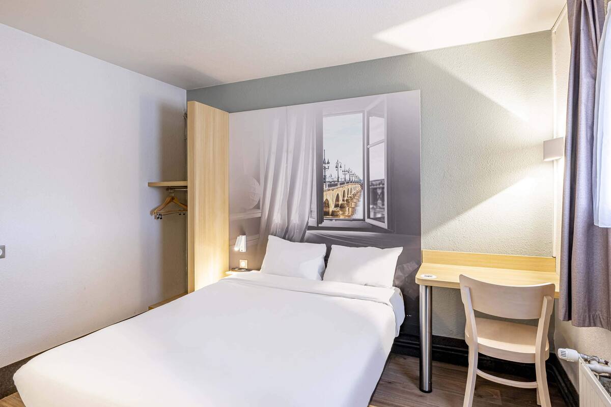 Ein Hotelzimmer mit Bett, Schreibtisch, Stuhl und einem großen Fenster mit Blick auf die Stadt.