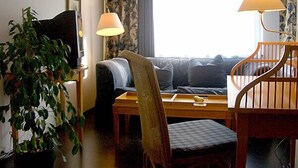 Ein Zimmer mit einer Couch, einem Sessel, einem Tisch und einer Lampe. Eine Pflanze und ein Fenster mit Vorhängen sind ebenfalls vorhanden.