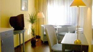 Ein kleines Hotelzimmer mit einem Einzelbett, einem Schreibtisch mit Lampe, einem Sessel, einem Fernseher und einem Blumentopf.