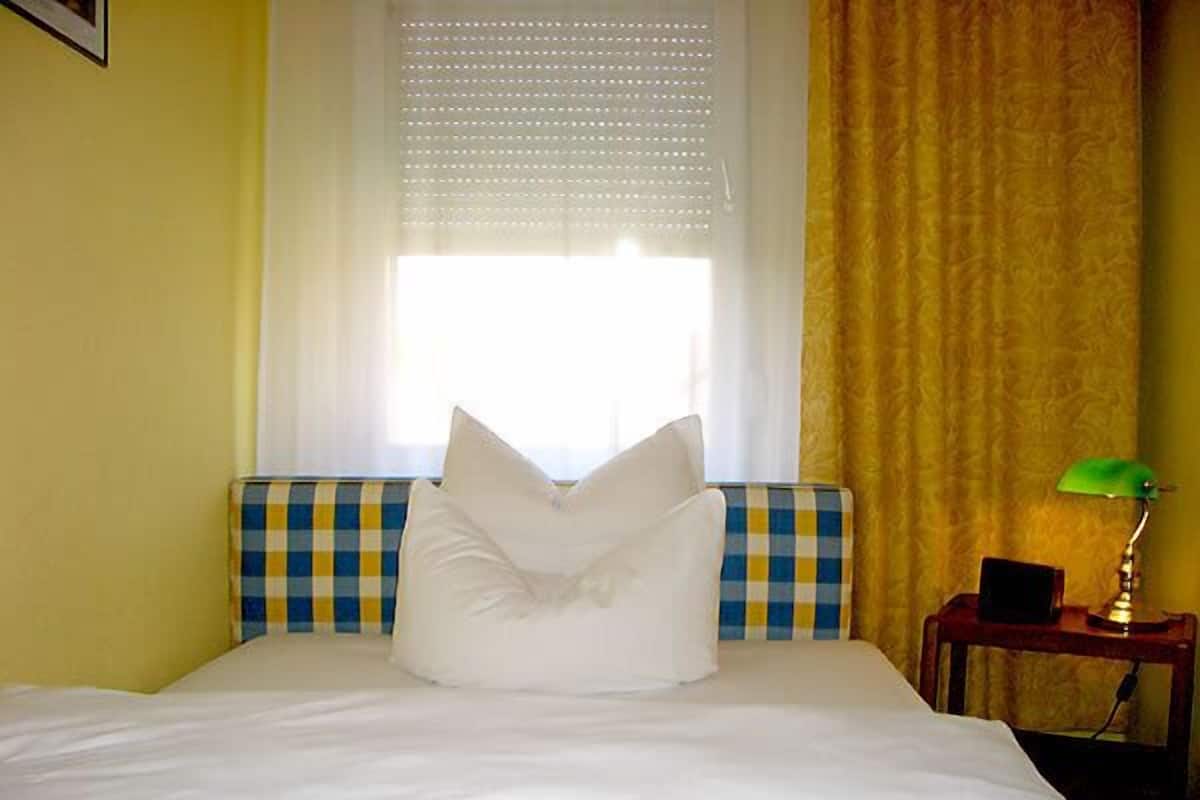 A bed with white bedding, a checkered headboard, and a pillow. There is a window with blinds and a curtain, a bedside table with a lamp, and a small black object on the table.
