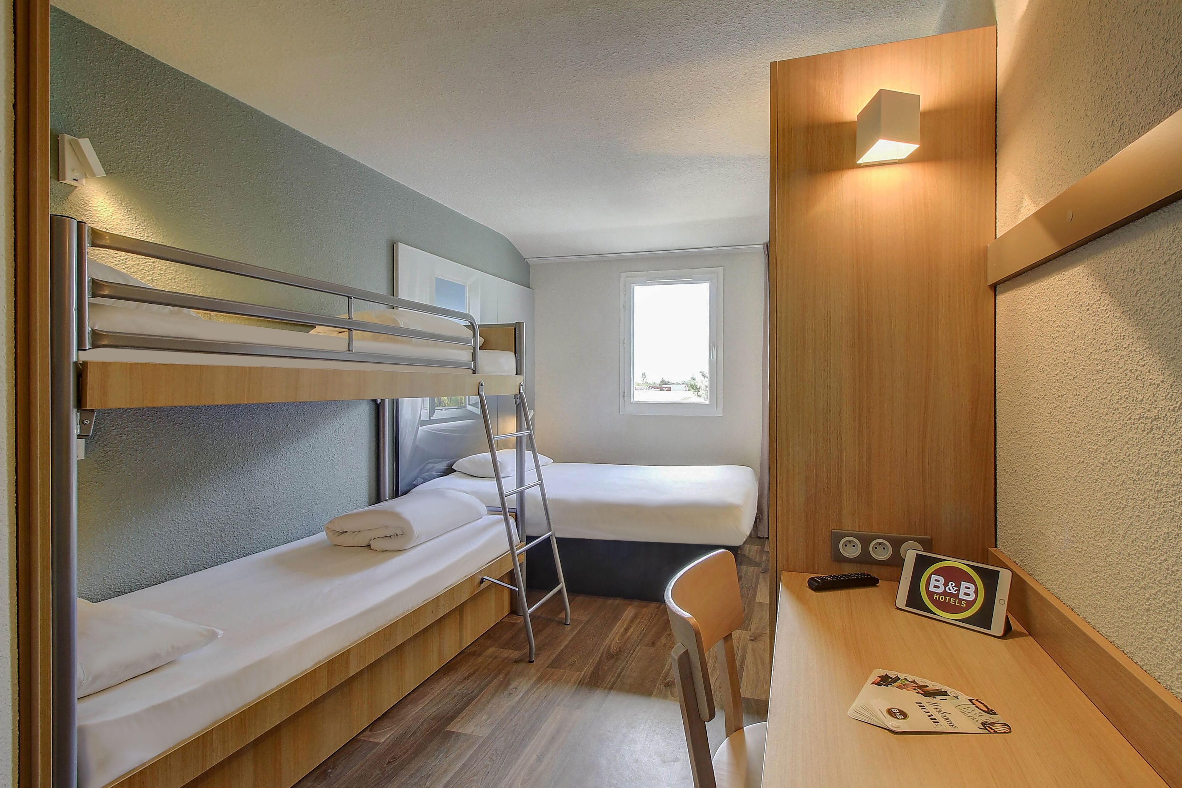 A compact room with bunk beds, a desk with a tablet, a chair, and a window.