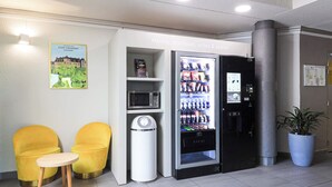 Een hotel lobby met een automaat, een magnetron, een prullenbak en twee gele stoelen.