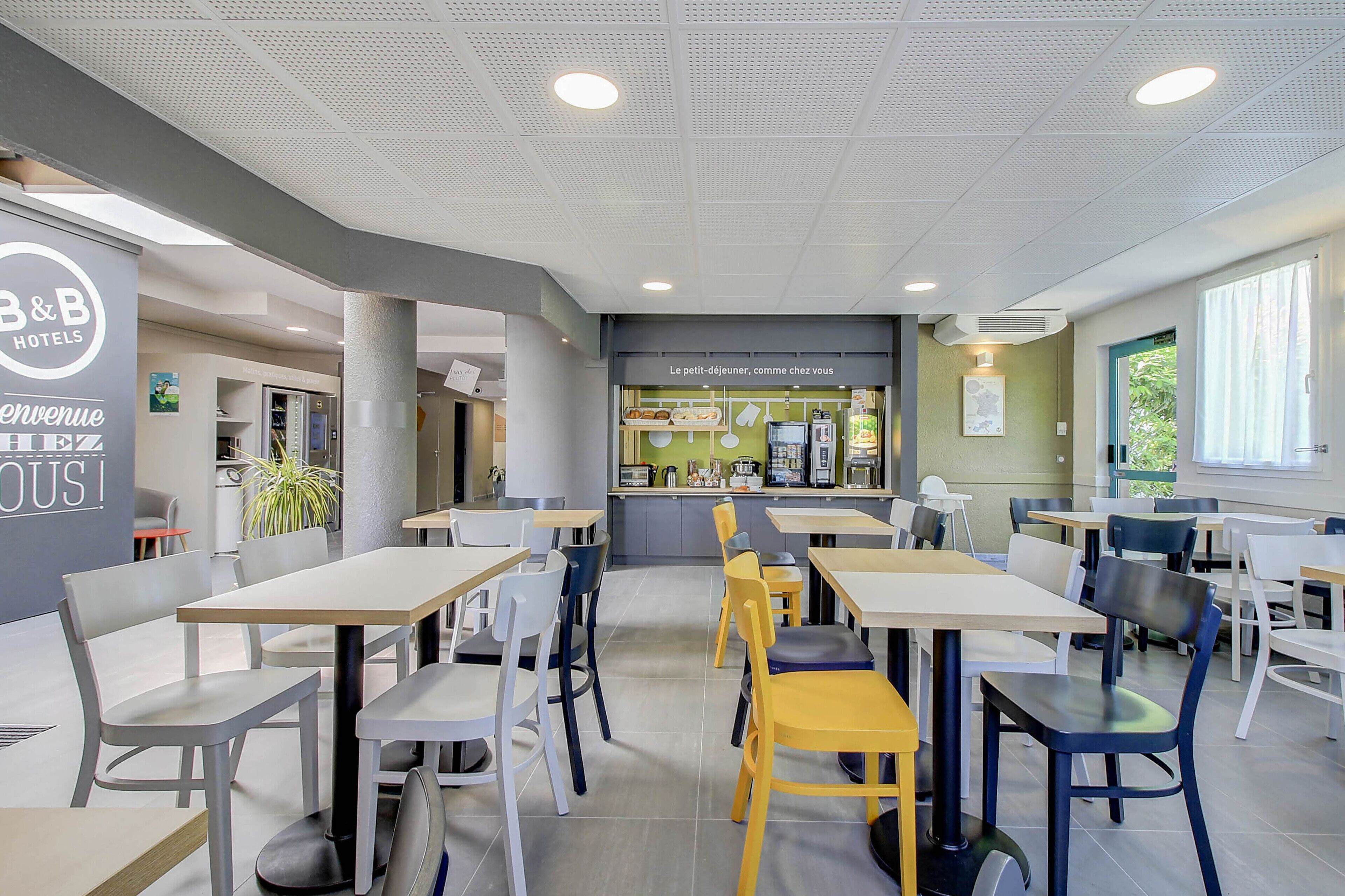 A modern hotel dining area with tables and chairs, a kitchenette, and a sign reading "B&B HOTEL".