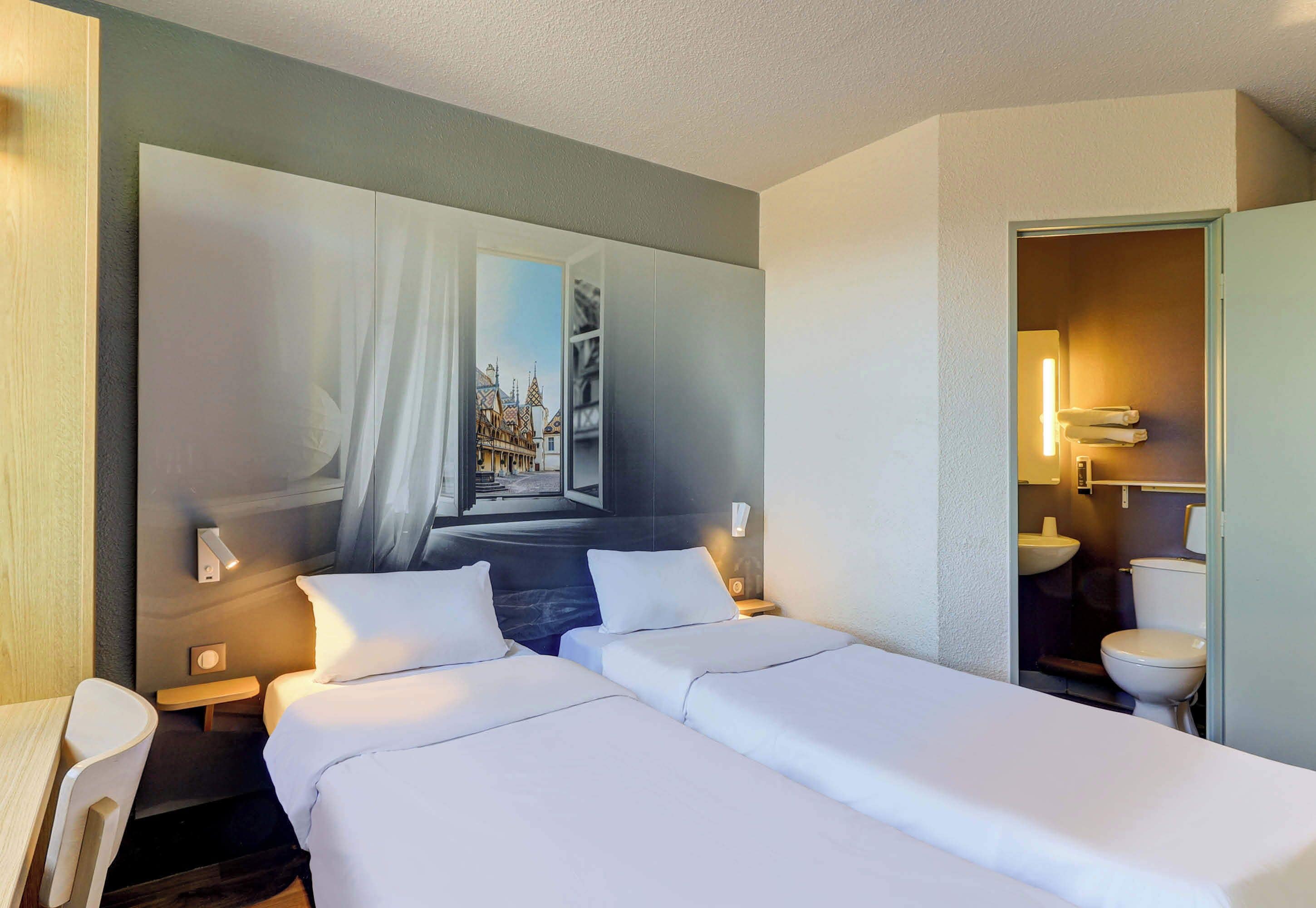 A hotel room with two beds, a large window with a view of a cityscape, a bedside table with a lamp, and a bathroom visible through an open door.