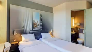 A hotel room with two beds, a large window with a view of a cityscape, a bedside table with a lamp, and a bathroom visible through an open door.