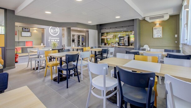 Een restaurantruimte in een hotel met tafels en stoelen, een receptiebalie en een keuken op de achtergrond.