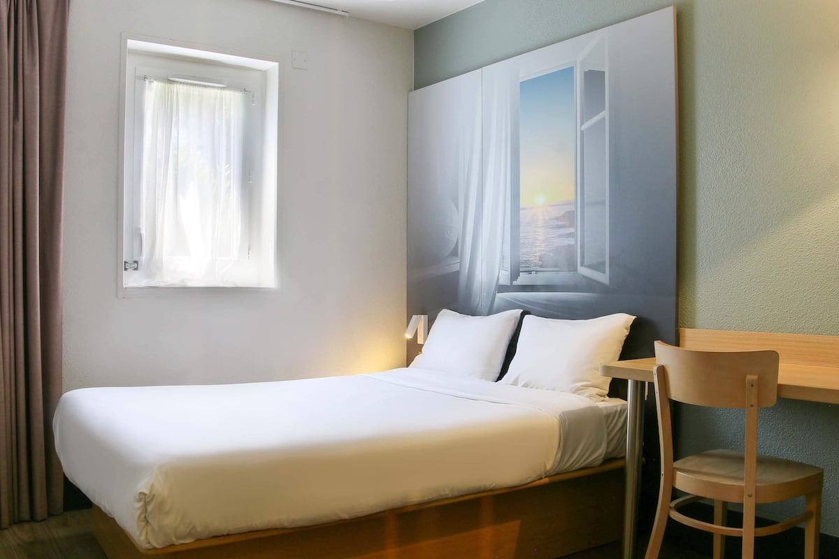 Une chambre d’hôtel avec un lit, une chaise, une fenêtre avec des rideaux et un tableau au mur.