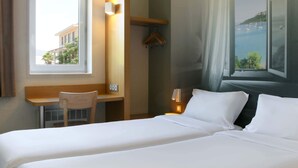 A hotel room with a bed, a desk, a chair, a coat rack, and a view of the sea. - B&B HOTEL Fréjus Roquebrune-sur-Argens (Roquebrune-sur-Argens)