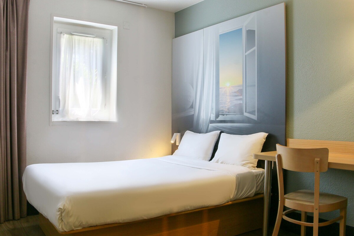 A hotel room with a bed, a chair, a window with curtains, and a painting on the wall.