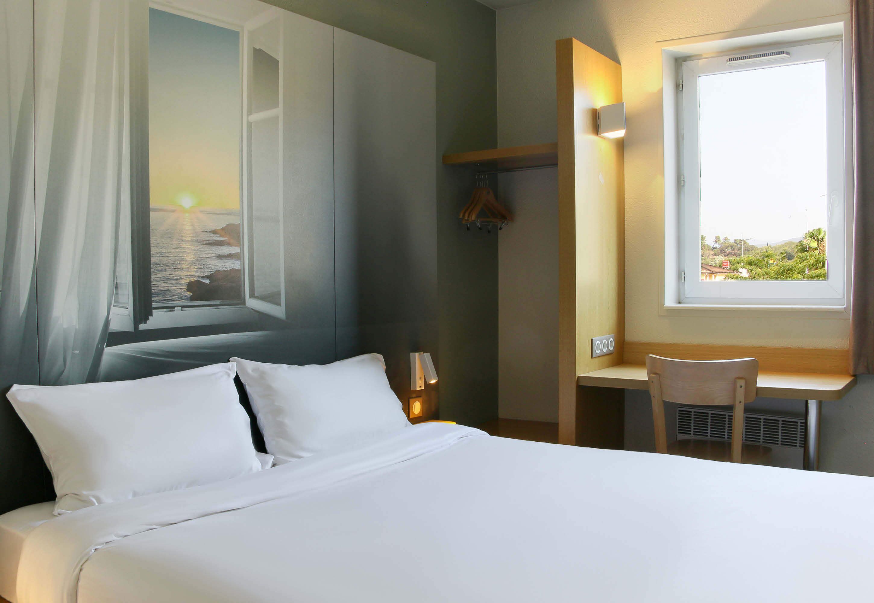 Una habitación de hotel con cama, escritorio, silla, ventana con vista al mar y perchero en la pared.