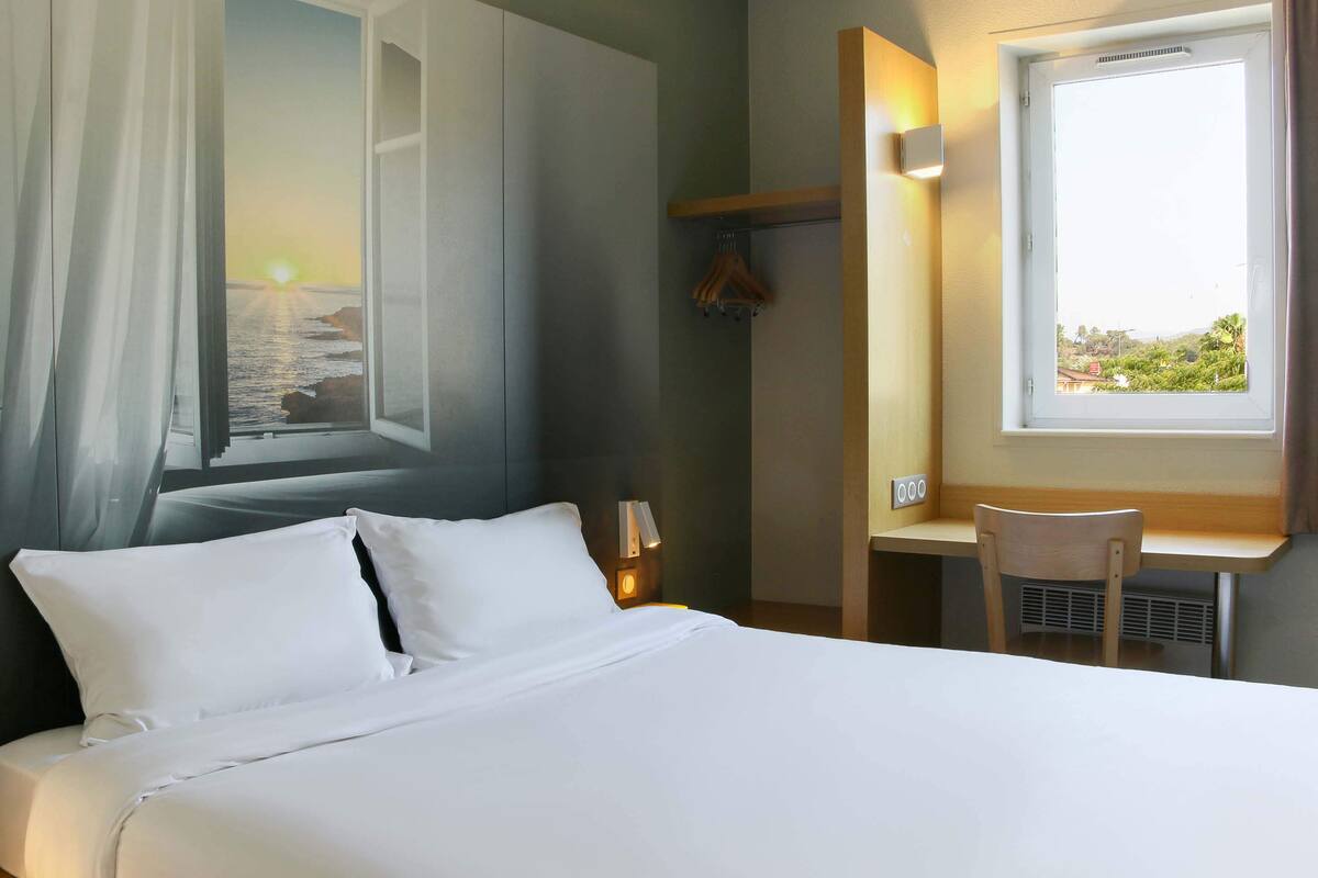 Une chambre d’hôtel comprenant un lit, un bureau, une chaise, une fenêtre donnant sur la mer et un porte-manteau fixé au mur.