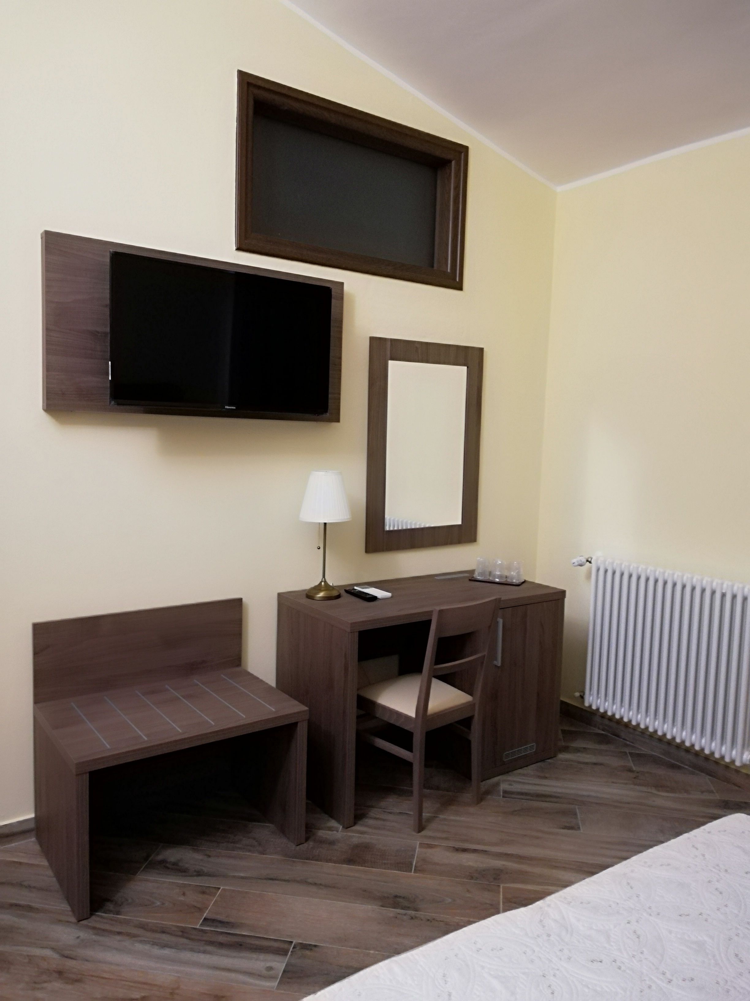 A hotel room with a bed, a desk with a chair, a TV, a mirror, a lamp, and a radiator.