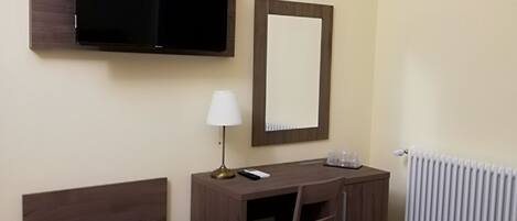 A hotel room with a bed, a desk with a chair, a TV, a mirror, a lamp, and a radiator.