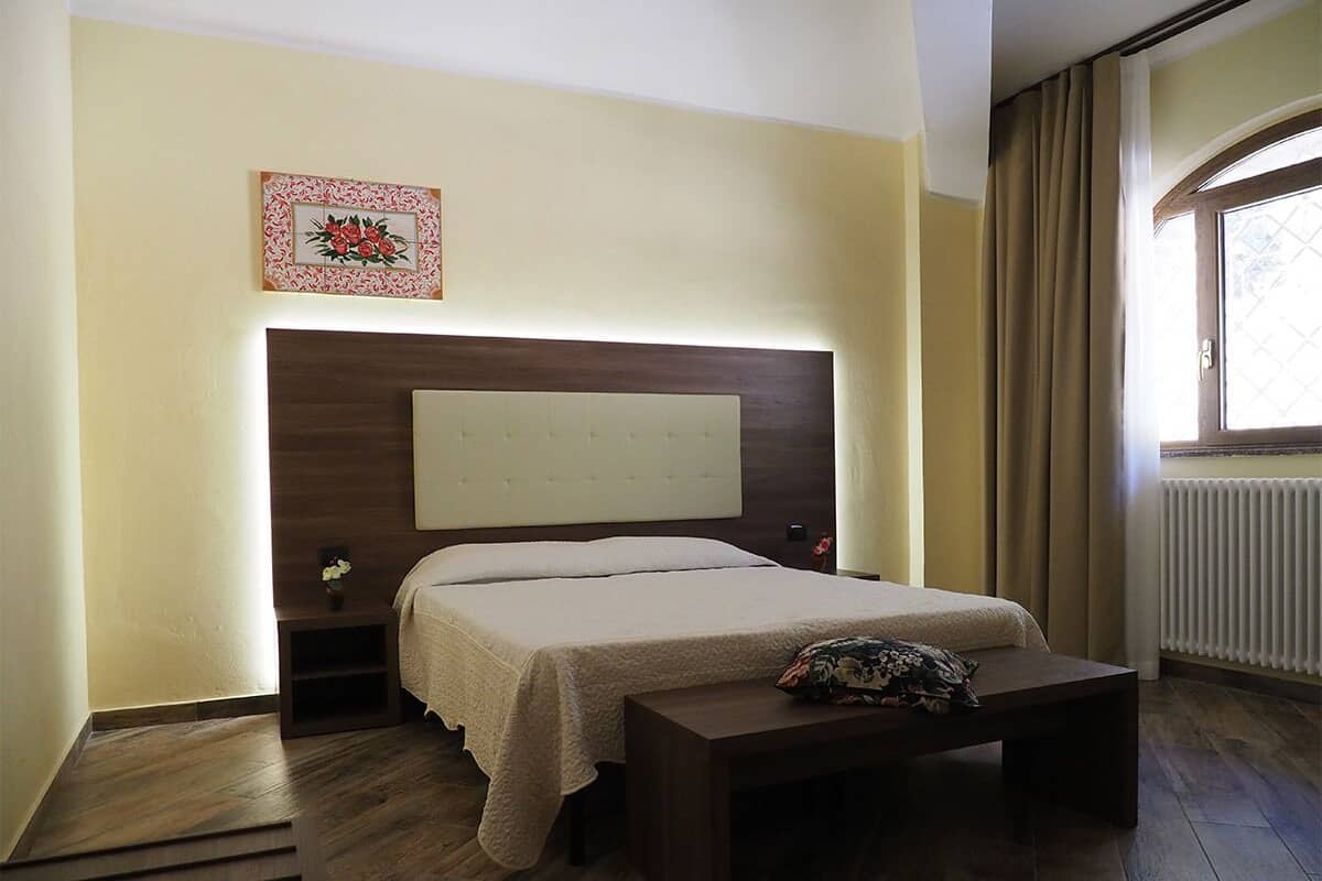 Une chambre à coucher comprenant un lit, des tables de chevet, une tête de lit, une fenêtre avec des rideaux et un radiateur.