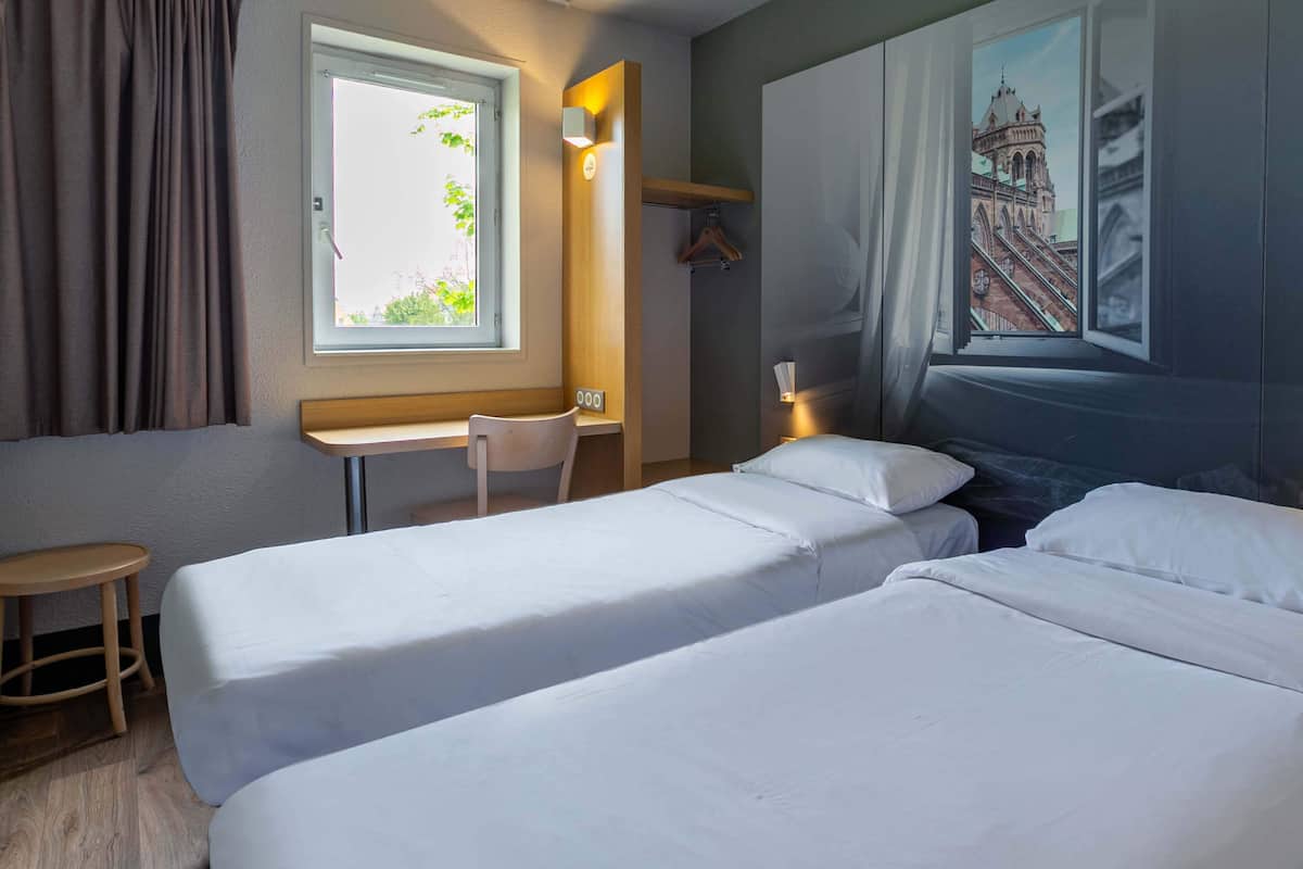 Une chambre d’hôtel avec deux lits simples, un petit bureau, une chaise, une fenêtre avec des rideaux et une lampe fixée au mur.
