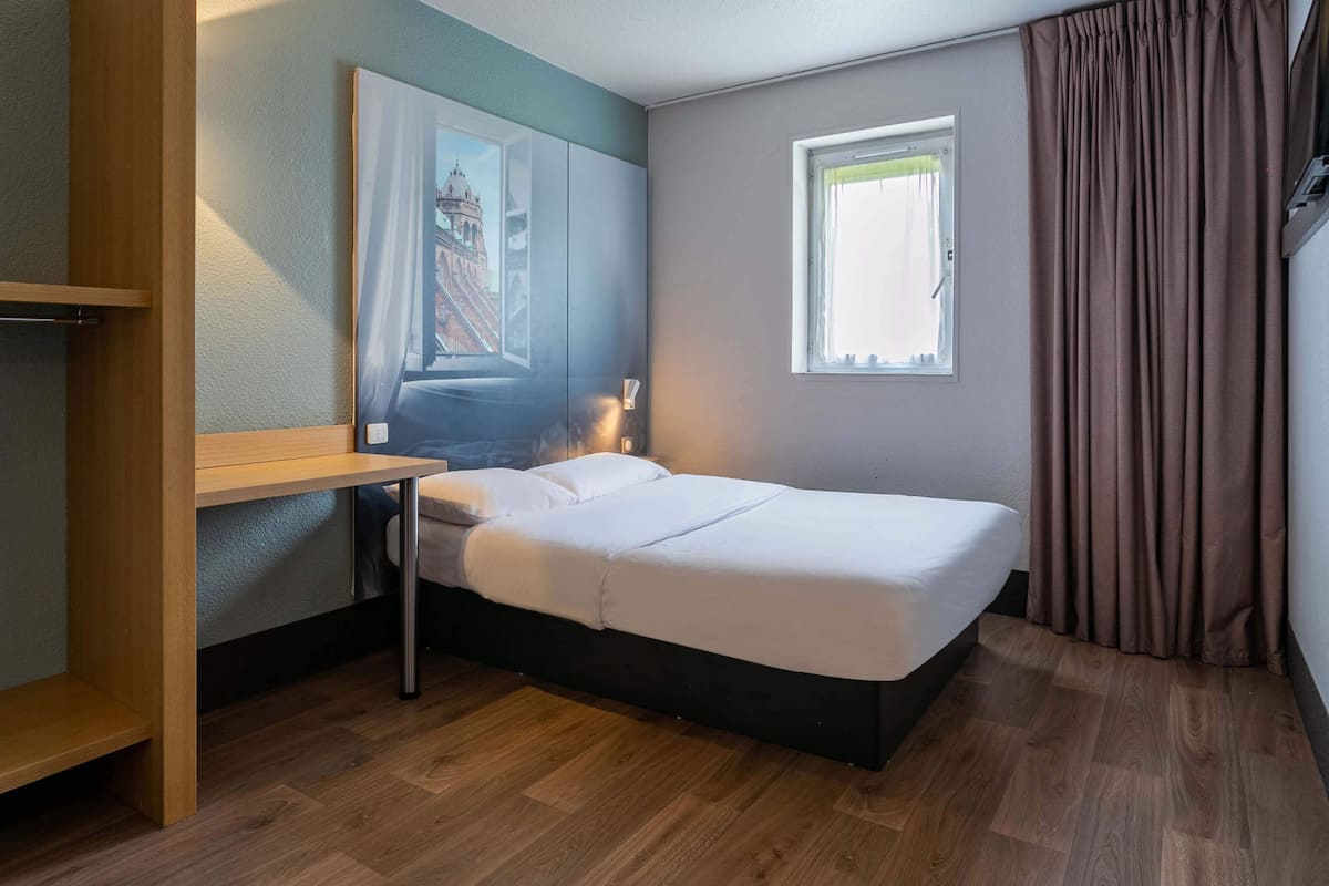 Une chambre d’hôtel moderne, équipée d’un lit, d’un bureau, d’une fenêtre et d’un tableau au mur.