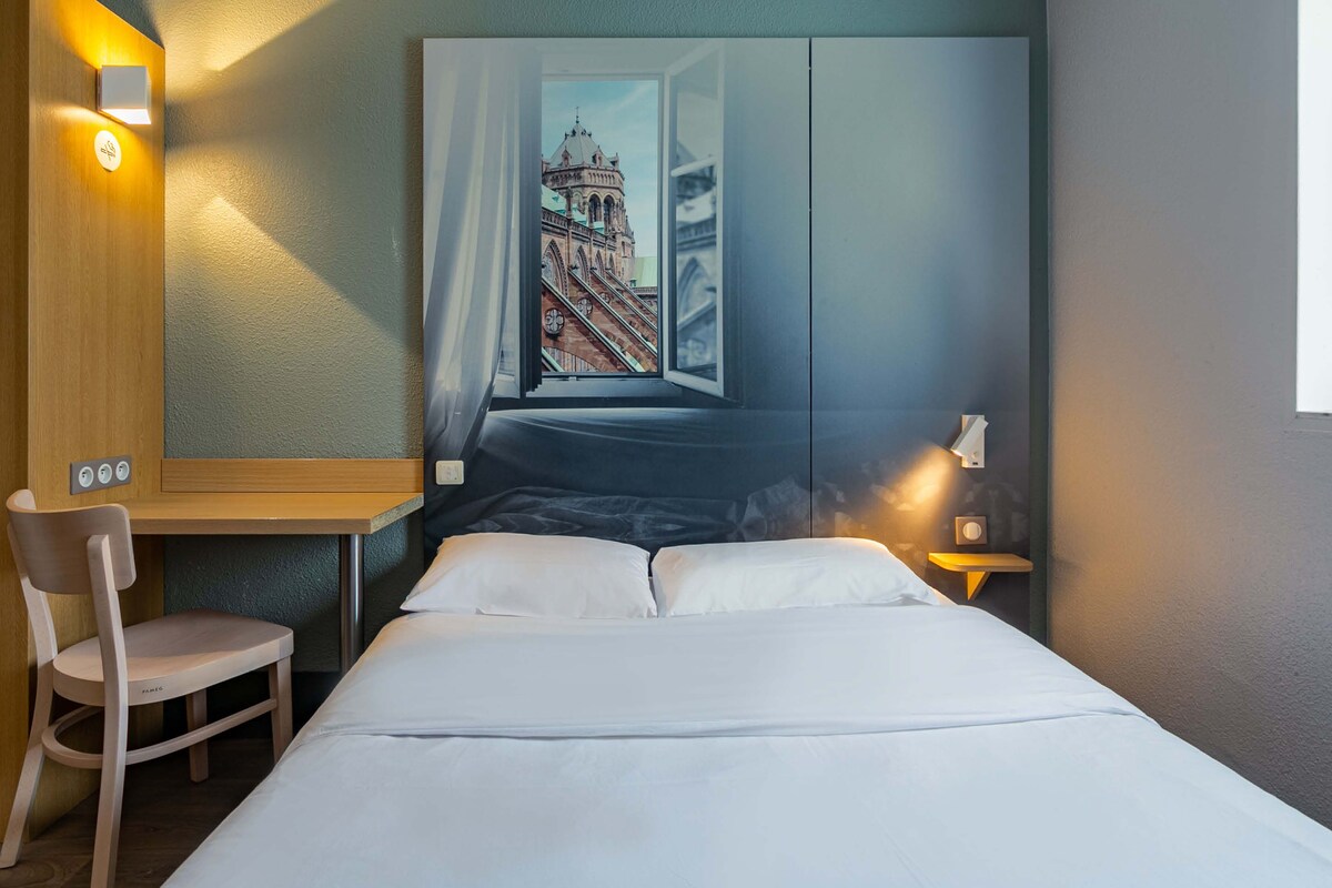 Ein Hotelzimmer mit Bett, Schreibtisch, Stuhl und einem großen Fenster mit Blick auf ein Gebäude.