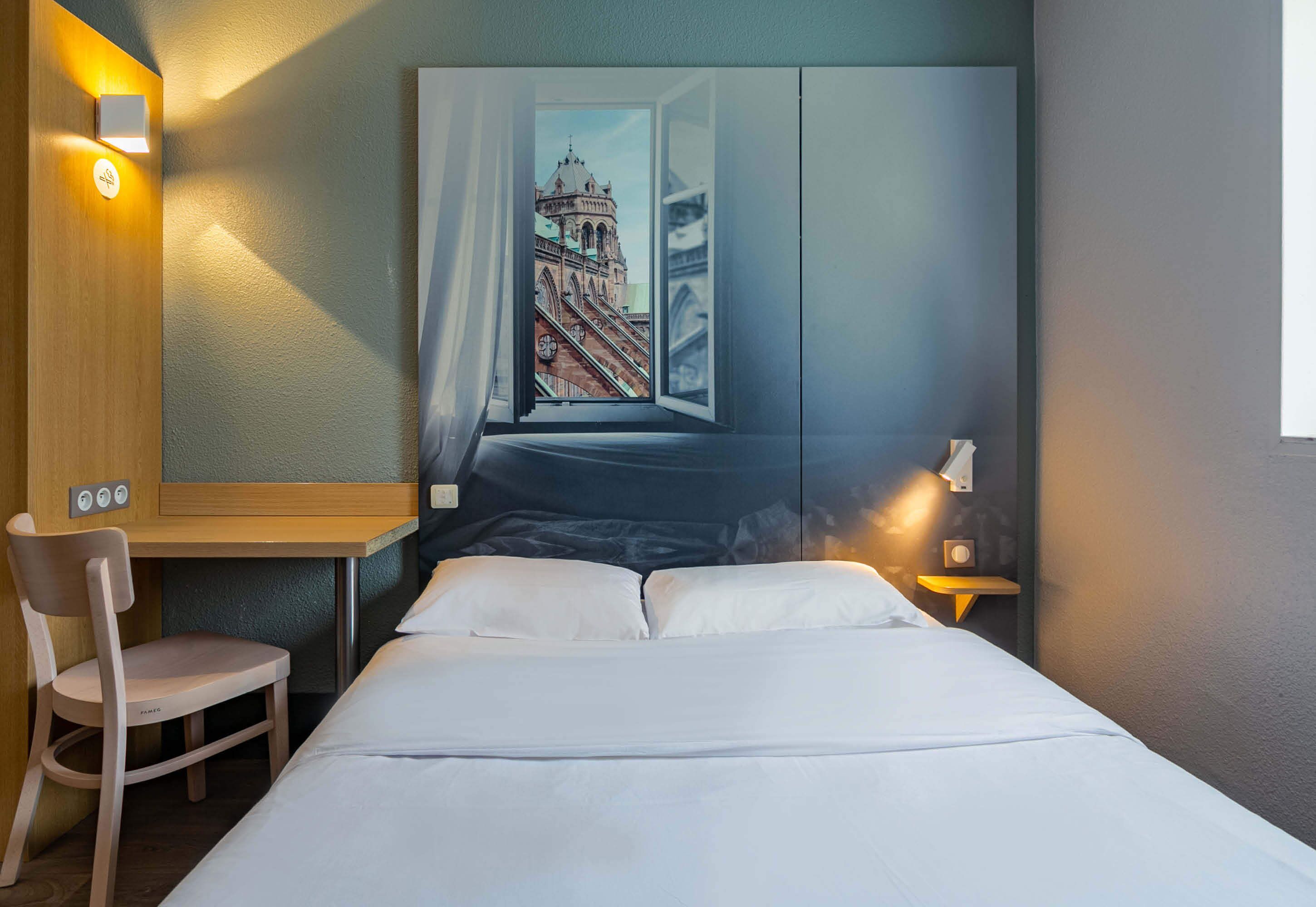 Une chambre d’hôtel avec un lit, un bureau, une chaise et une grande fenêtre donnant sur un immeuble.