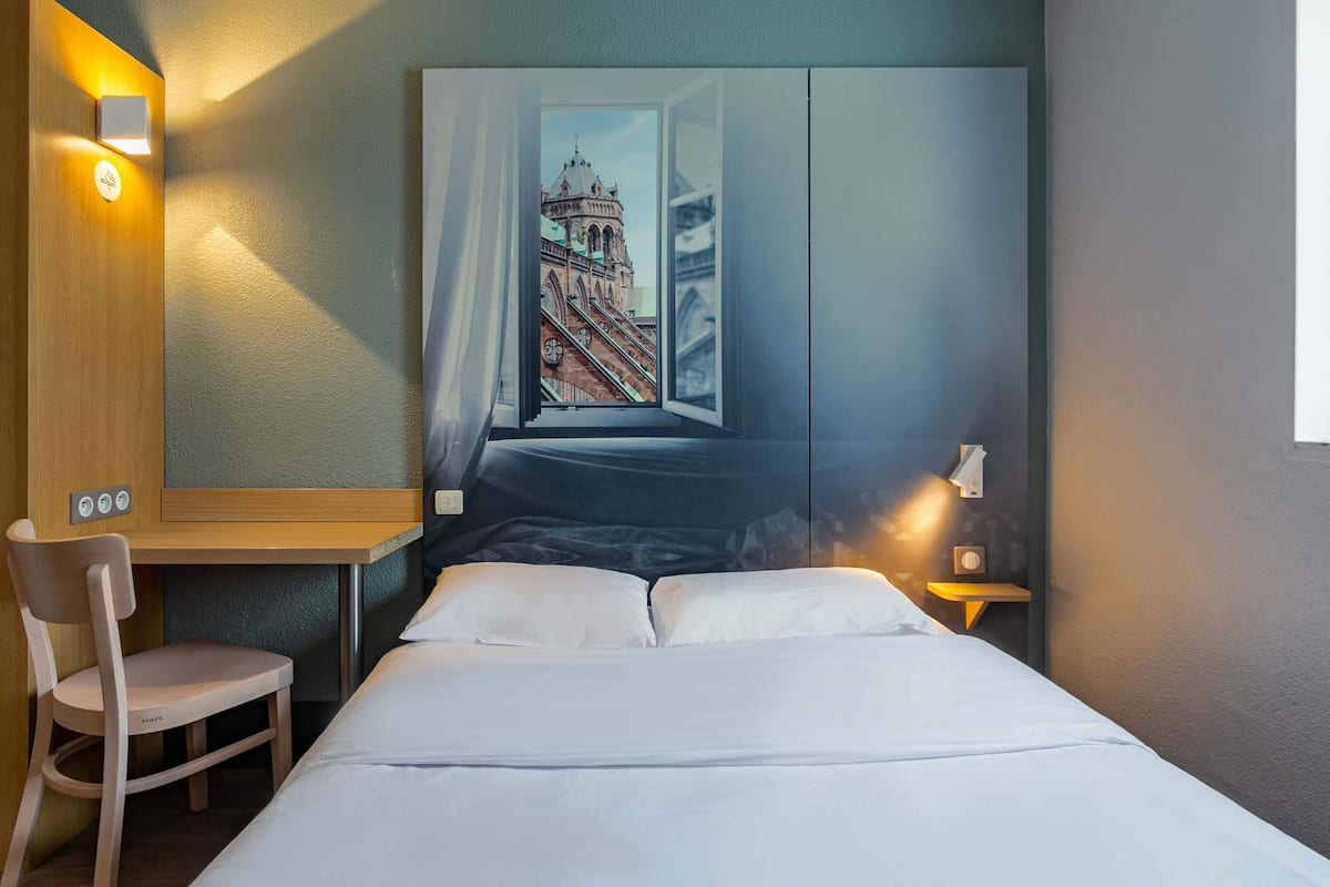 Une chambre d’hôtel avec un lit, un bureau, une chaise et une grande fenêtre donnant sur un immeuble.
