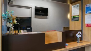 A reception desk with a flat-screen TV, a wooden counter, and a vase with flowers. - B&B HOTEL Strasbourg Nord Artisans (Vendenheim)