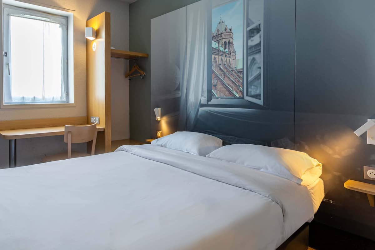 Une chambre d’hôtel avec un lit, un bureau et une chaise. Il y a un tableau représentant un bâtiment accroché au mur.