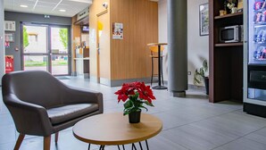 A modern lobby with a leather chair, a round wooden table with a potted plant, and a vending machine. - B&B HOTEL Strasbourg Nord Artisans (Vendenheim)