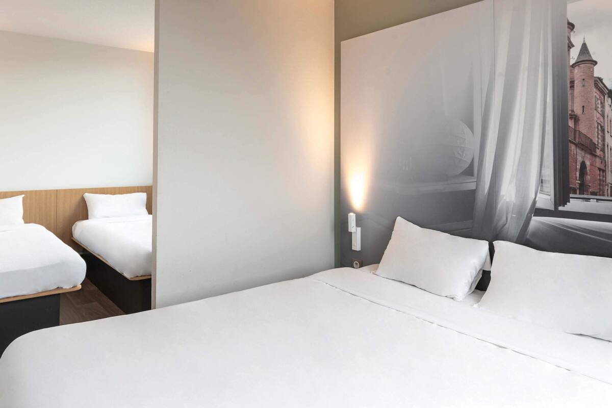 Habitación de hotel con una cama grande, dos camas más pequeñas, una lámpara fijada a la pared y vista a un edificio a través de una ventana.