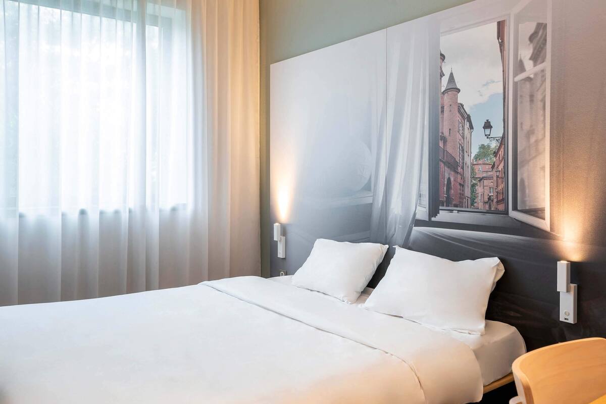 Habitación de hotel con una cama grande, ropa blanca, una ventana con cortinas traslúcidas y un cuadro de un paisaje urbano en la pared.