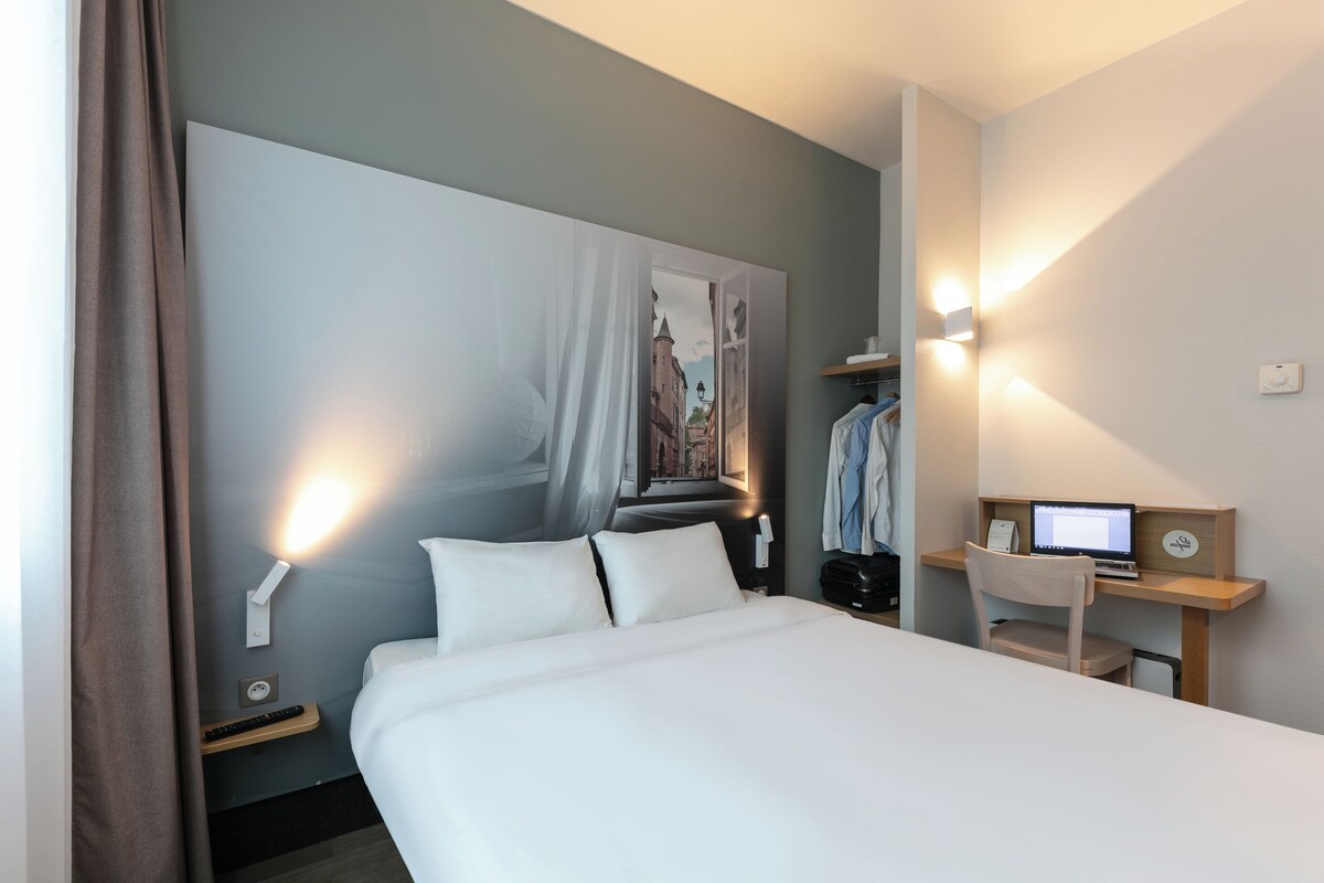 A hotel room with a bed, a desk with a computer, a chair, a coat rack, and a wall-mounted light.