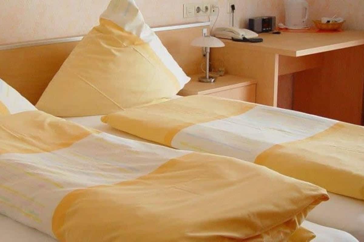 Quarto de hotel com duas camas, cada uma com roupa de cama e almofadas amarelas. Apresenta uma mesa de cabeceira de madeira com uma lâmpada e um telefone.