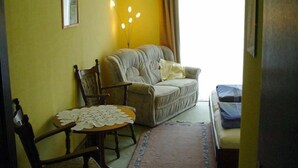 Una habitación con un sofá, una silla, una mesa pequeña con mantel de encaje y una cama con manta azul.